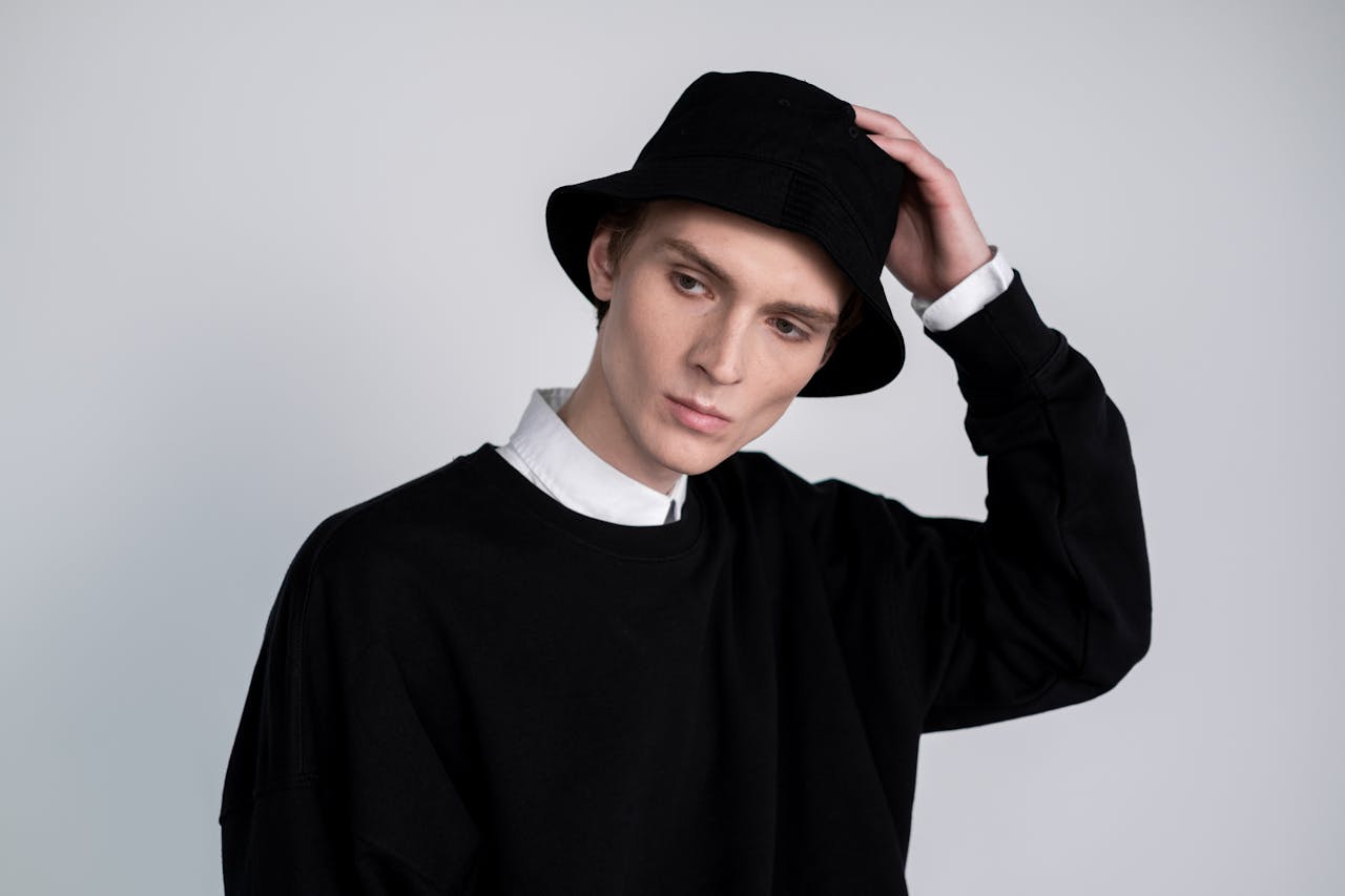 Male model in black clothing and bucket hat posing thoughtfully in a studio.